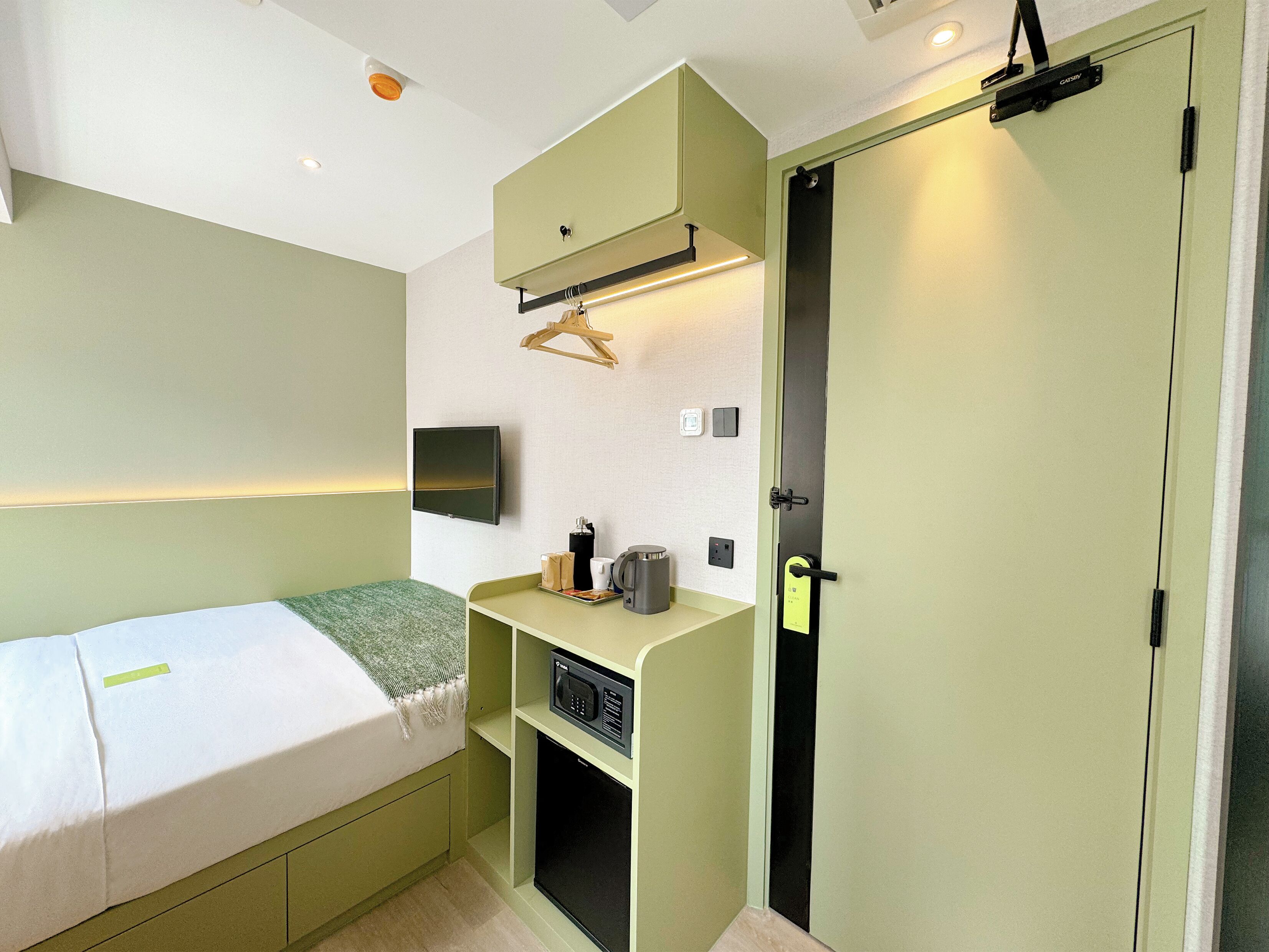 A compact hotel room with a bed, a TV mounted on the wall, a microwave, and a small shelf with toiletries.