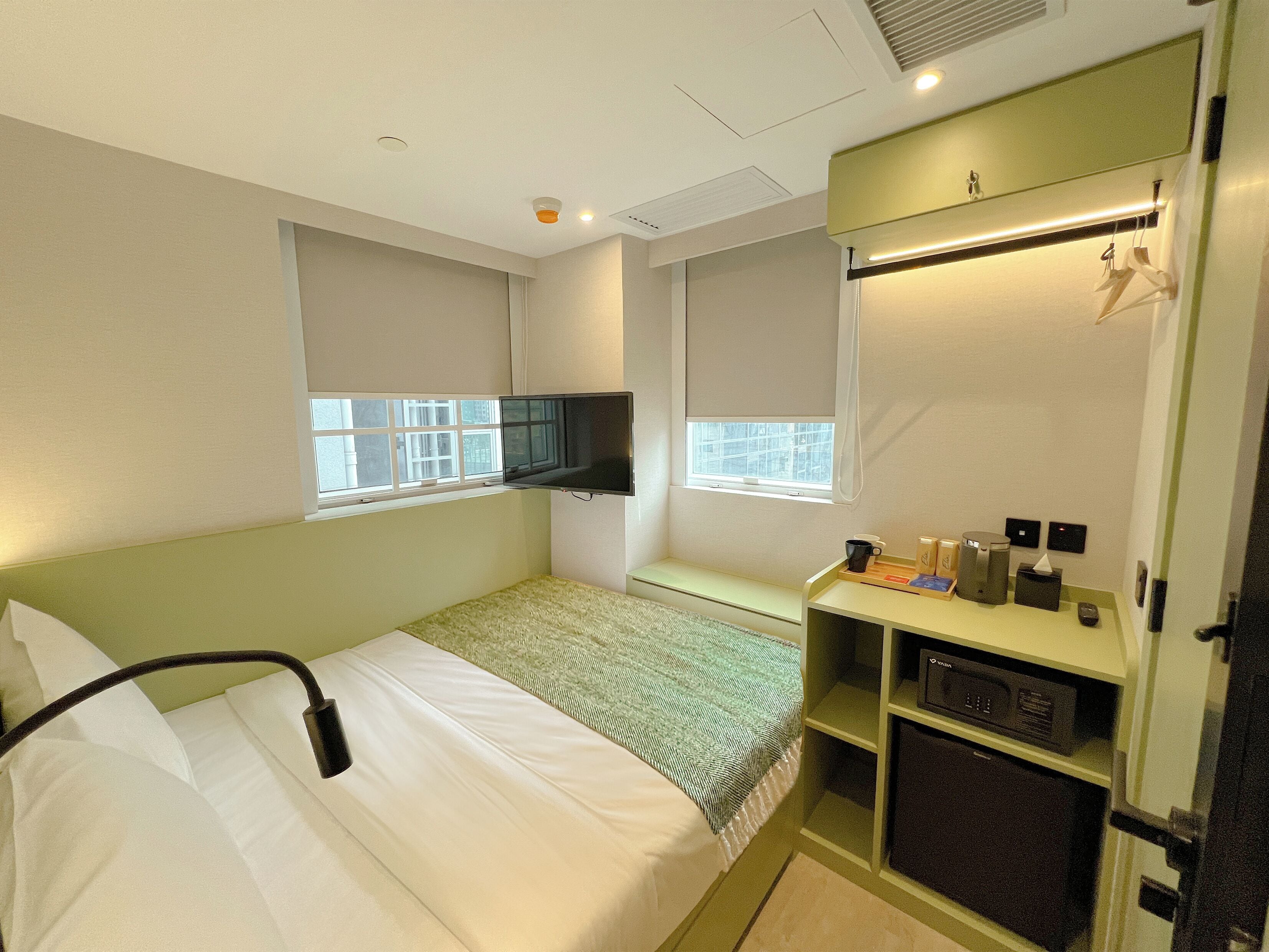 Een compacte hotelkamer met een enkel bed, een aan de muur bevestigde televisie, een klein zithoekje en een kitchenette met een magnetron.