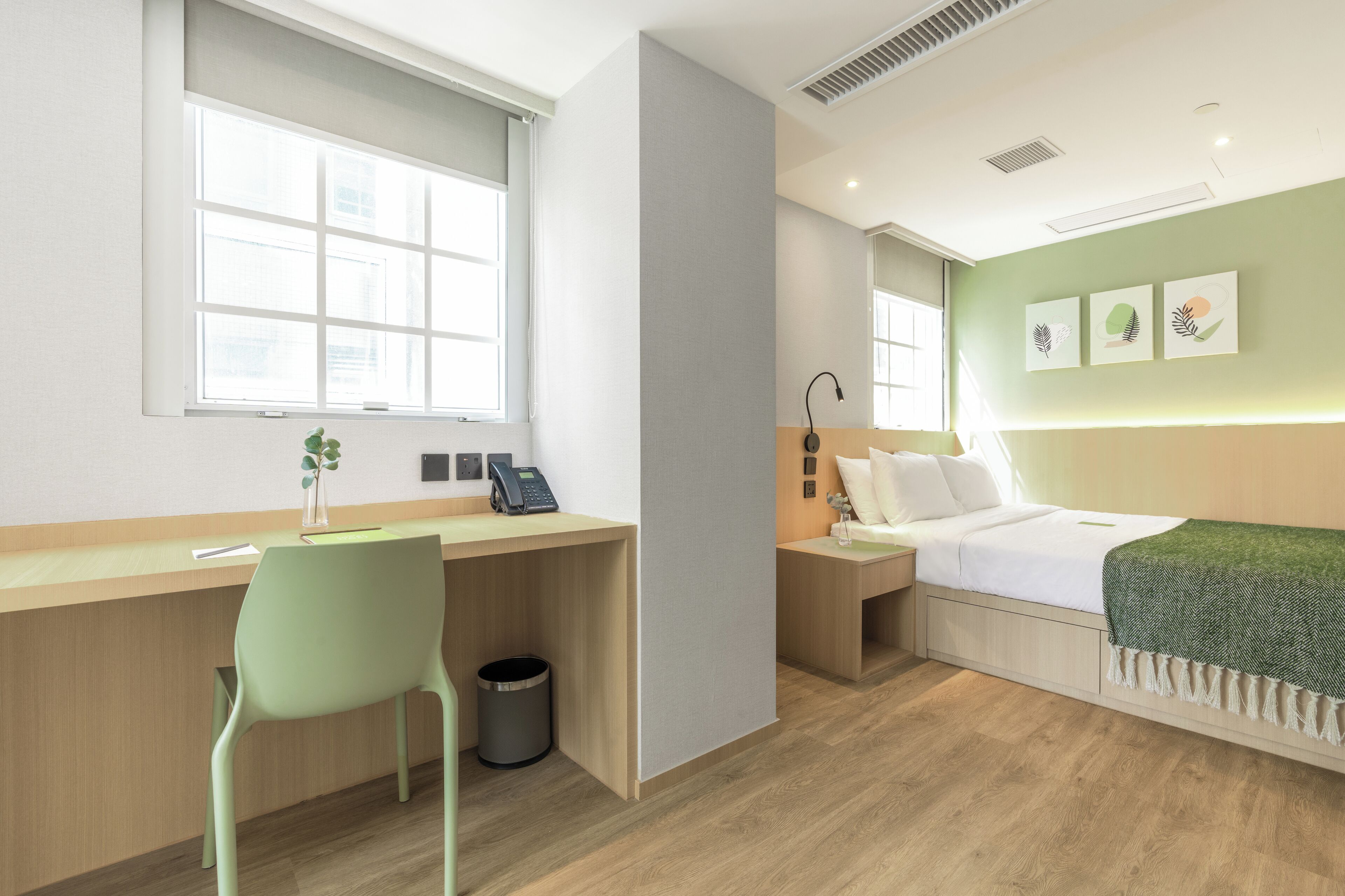 Une chambre d’hôtel moderne avec un bureau en bois, une chaise verte, un lit recouvert d’un plaid vert et une table de chevet avec une lampe.