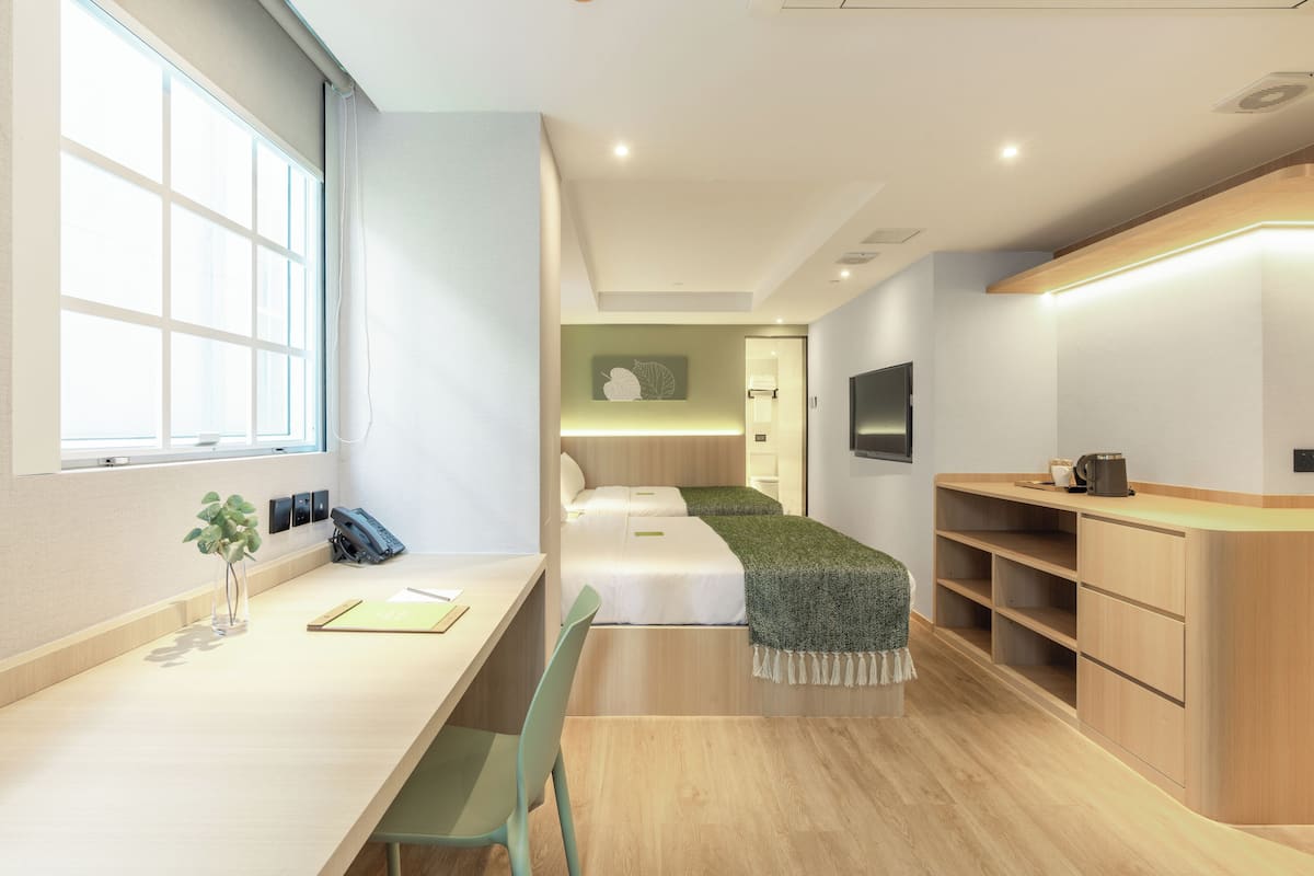 A modern hotel room with a wooden desk, a chair, a bed with a green blanket, a TV, and a bedside table with a coffee maker.