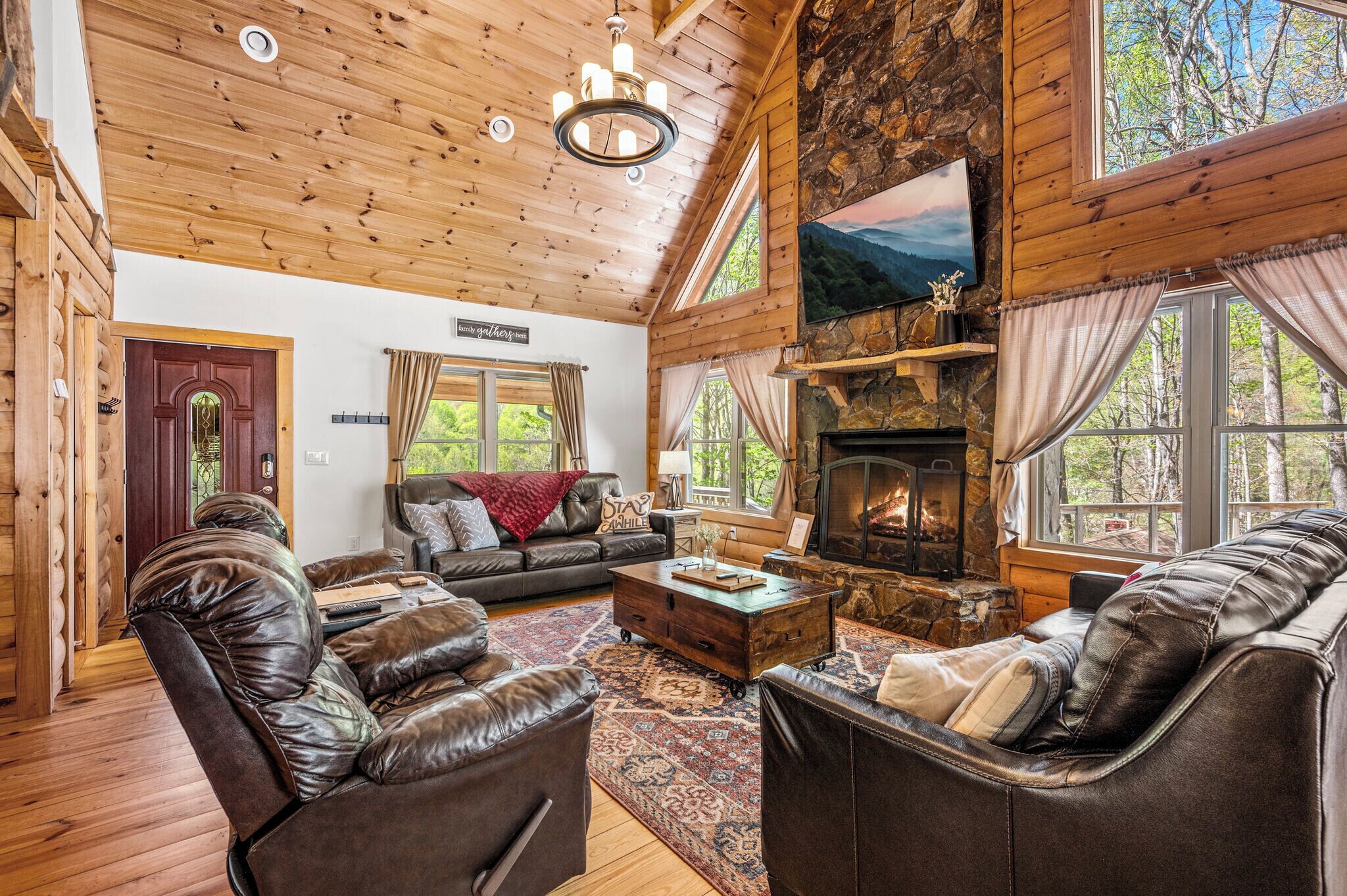 A cozy log cabin interior with a fireplace, leather sofas, and a large window offering a view of the forest.