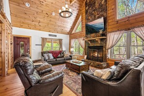A cozy log cabin interior with a fireplace, leather sofas, and a large window offering a view of the forest. - Cataloochee Chalet Mountain Views/Hot Tub/Fire Pit (Maggie Valley)