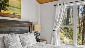 A bedroom with a wooden headboard, a bed with patterned bedding, a nightstand with a lamp, and a window with a forest view. - Cataloochee Chalet Mountain Views/Hot Tub/Fire Pit (Maggie Valley)