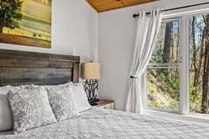 A bedroom with a wooden headboard, a bed with patterned bedding, a nightstand with a lamp, and a window with a forest view.