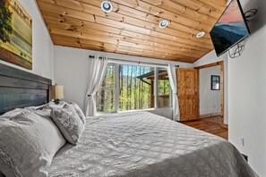 A bedroom with a large bed, a wooden ceiling, and a view of a forest through a window. - Cataloochee Chalet Mountain Views/Hot Tub/Fire Pit (Maggie Valley)