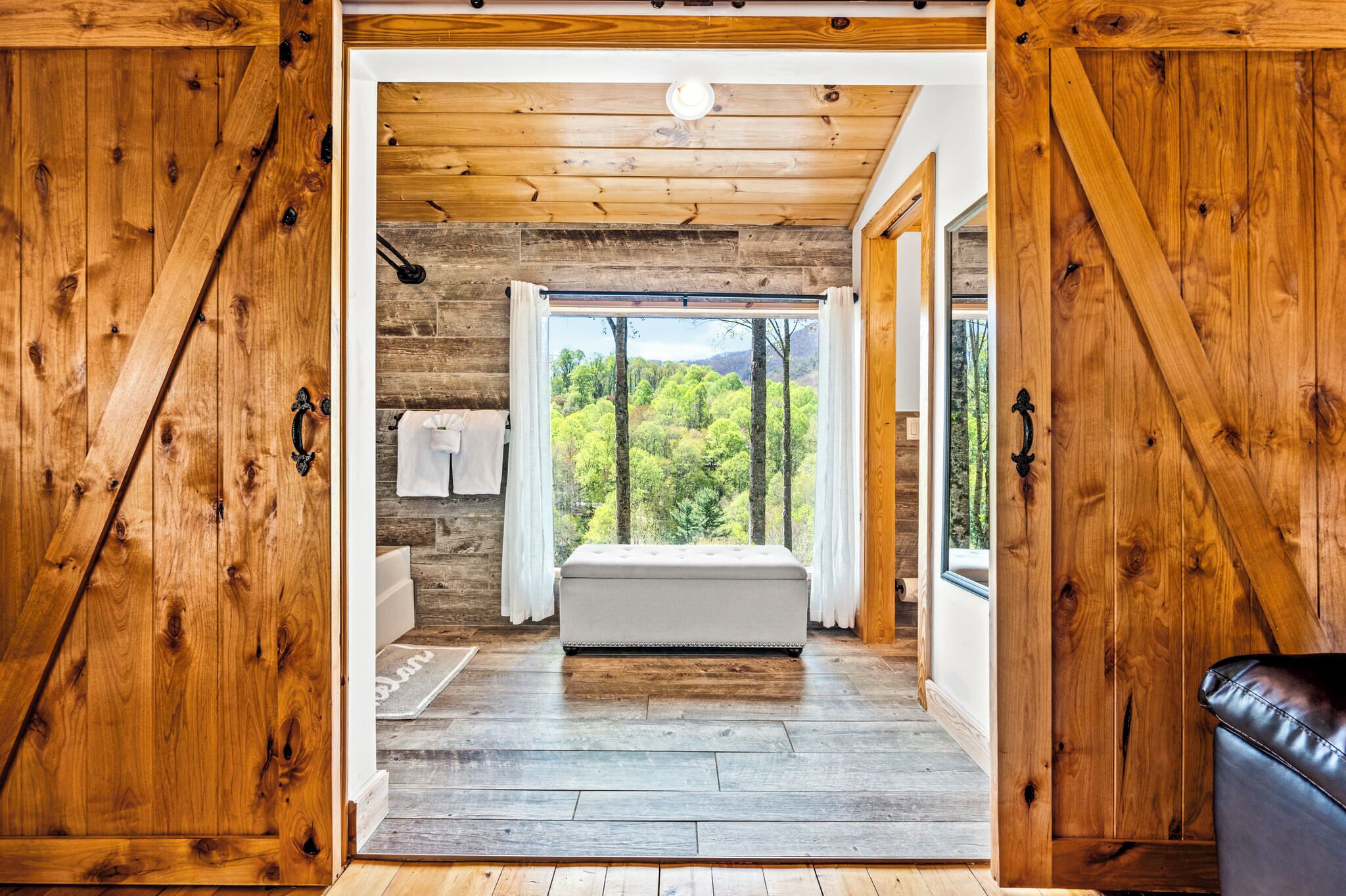 A wooden cabin interior with a bed, a bench, and a view of a forest through a window.