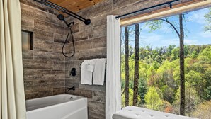 A modern bathroom with a bathtub, a shower, and a large window offering a forest view. - Cataloochee Chalet Mountain Views/Hot Tub/Fire Pit (Maggie Valley)