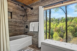 A modern bathroom with a bathtub, a shower, and a large window offering a forest view.
