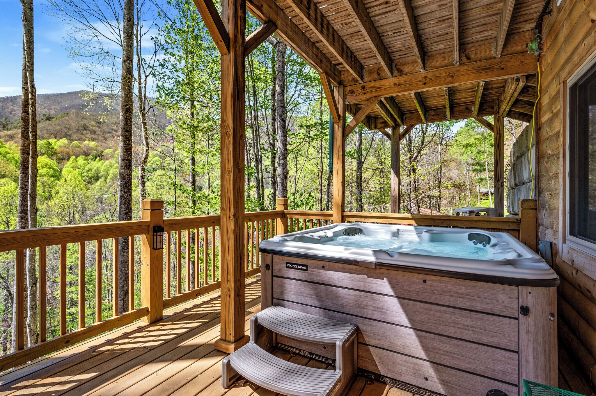 A wooden deck with a hot tub, wooden railings, and a view of a forested area.