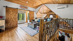 A rustic cabin interior with wooden floors, a loft bedroom with a sloped ceiling, a large sliding door, and a cozy seating area with a sofa and cushions. - Cataloochee Chalet Mountain Views/Hot Tub/Fire Pit (Maggie Valley)