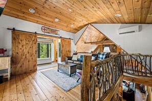 A rustic cabin interior with wooden floors, a loft bedroom with a sloped ceiling, a large sliding door, and a cozy seating area with a sofa and cushions.