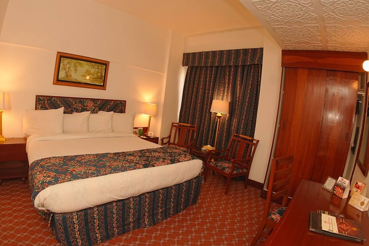 A hotel room with a bed, two bedside tables, a chair, a desk, a mirror, a door, and a painting on the wall.