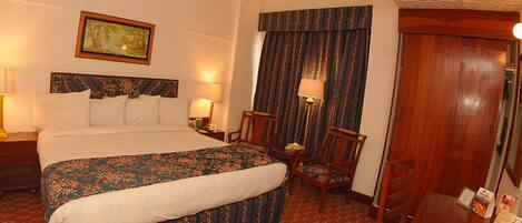 A hotel room with a bed, two bedside tables, a chair, a desk, a mirror, a door, and a painting on the wall.