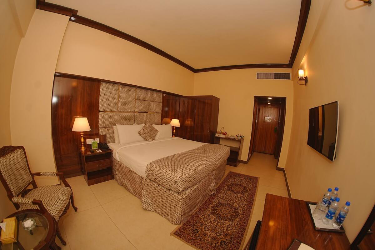 A hotel room with a bed, two bedside tables with lamps, a chair, a desk with water bottles, and a TV mounted on the wall.