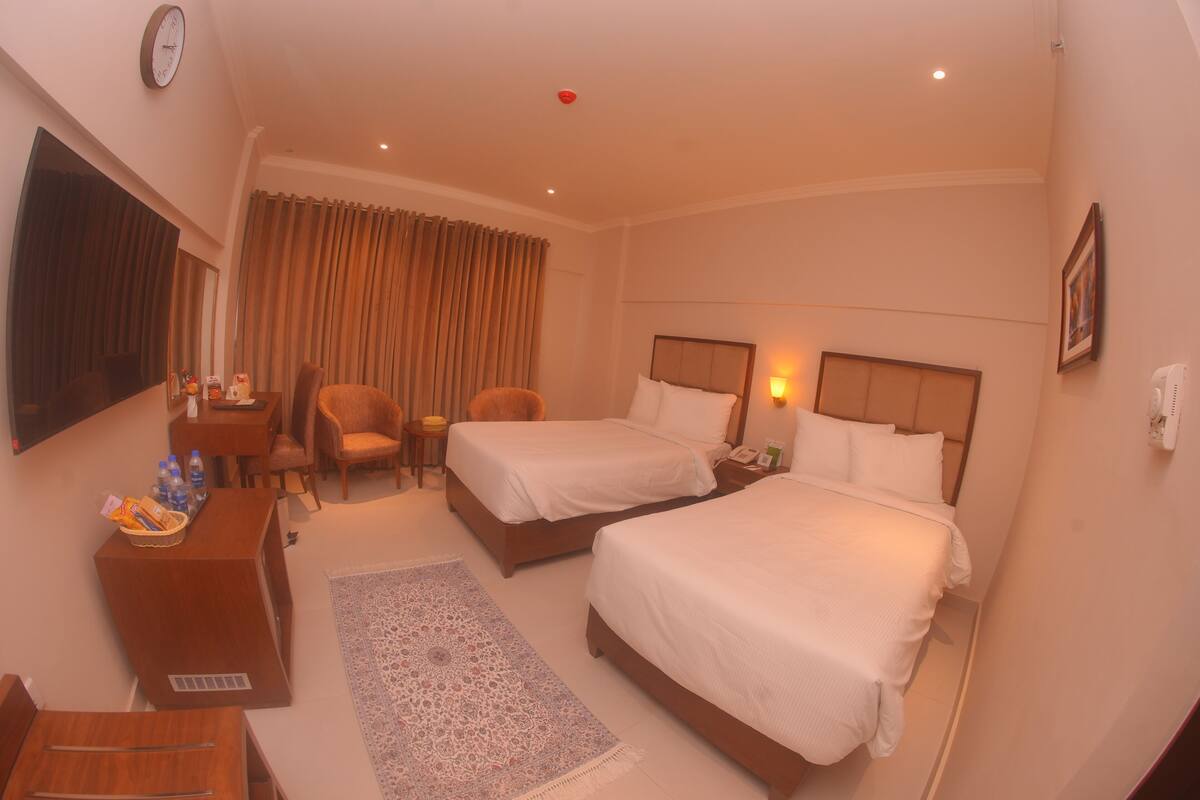 A hotel room with two beds, a desk, a chair, a TV, and a clock on the wall.