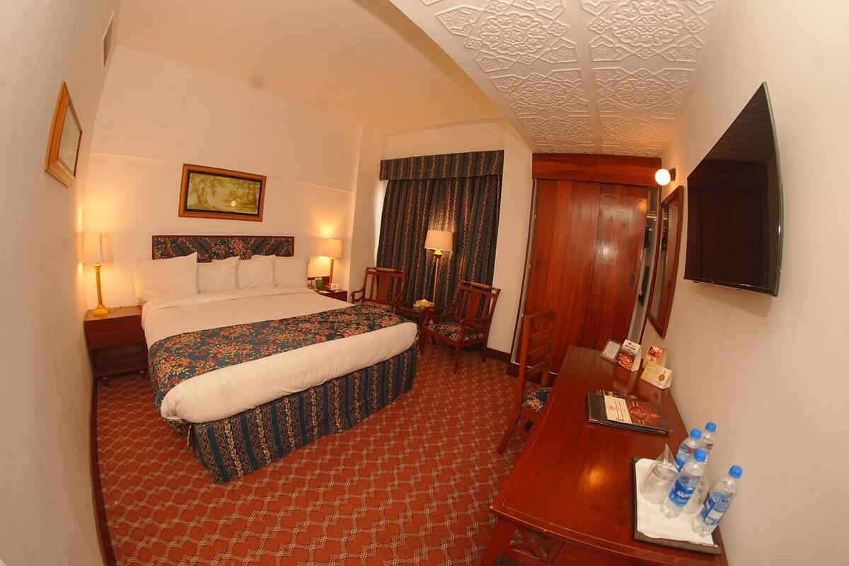 A hotel room with a bed, two bedside tables, a desk with chairs, a flat-screen TV, and a patterned carpet.
