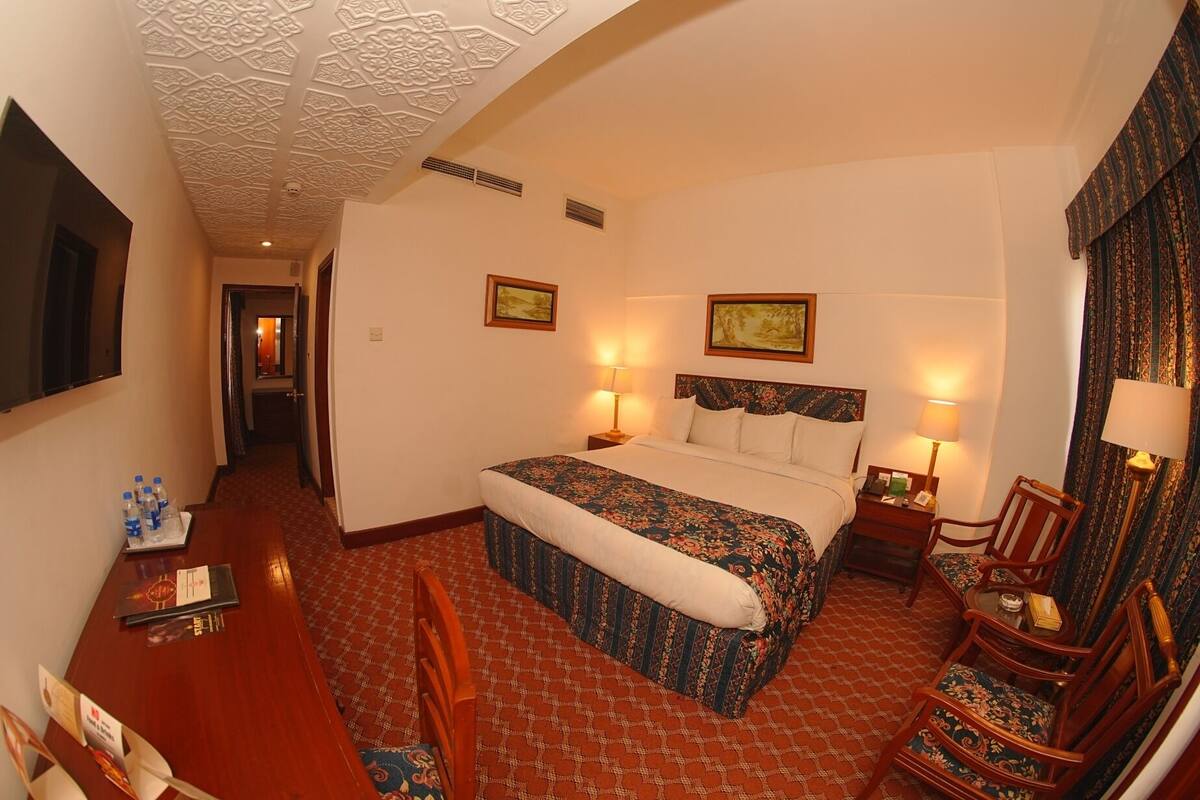 A hotel room with a bed, two bedside tables with lamps, a chair, a desk with a chair, and a patterned carpet.