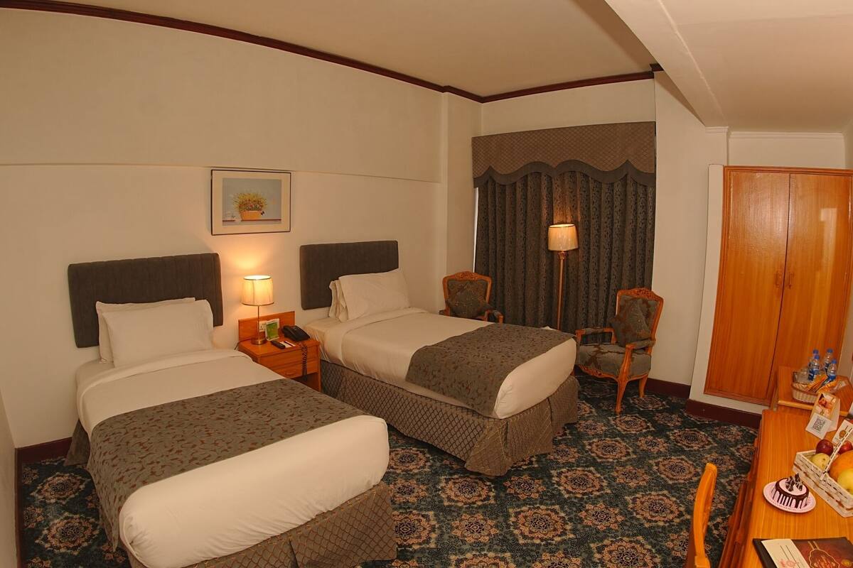 A hotel room with two beds, a desk with a chair, a nightstand, a lamp, a wardrobe, and a patterned carpet.