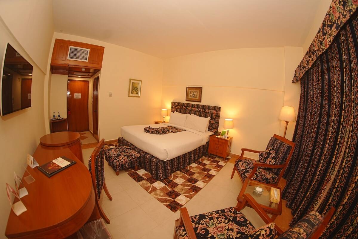 A hotel room with a bed, bedside tables, a desk, a chair, and a patterned curtain.