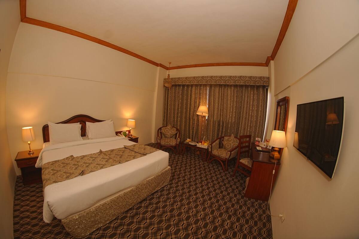 A hotel room with a large bed, two bedside tables with lamps, a small table with chairs, and a large window with curtains.