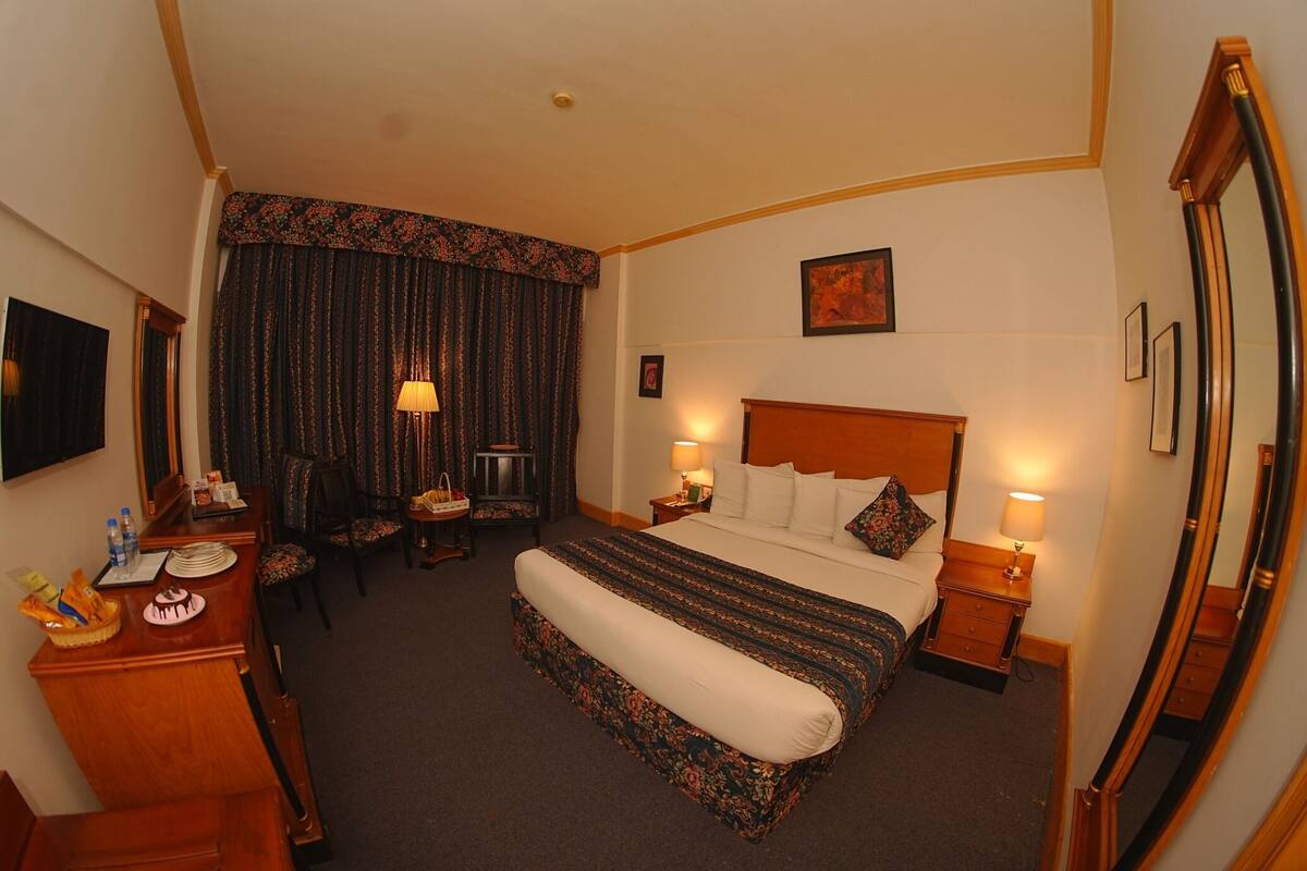 A hotel room with a bed, bedside tables, a desk with a chair, a dining table, and a large mirror.