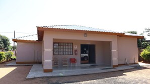 A small building with a metal roof, a door, and a window. There are two chairs outside and a fire extinguisher mounted on the wall. - Amariah Guesthouse Newstance (Kasane)