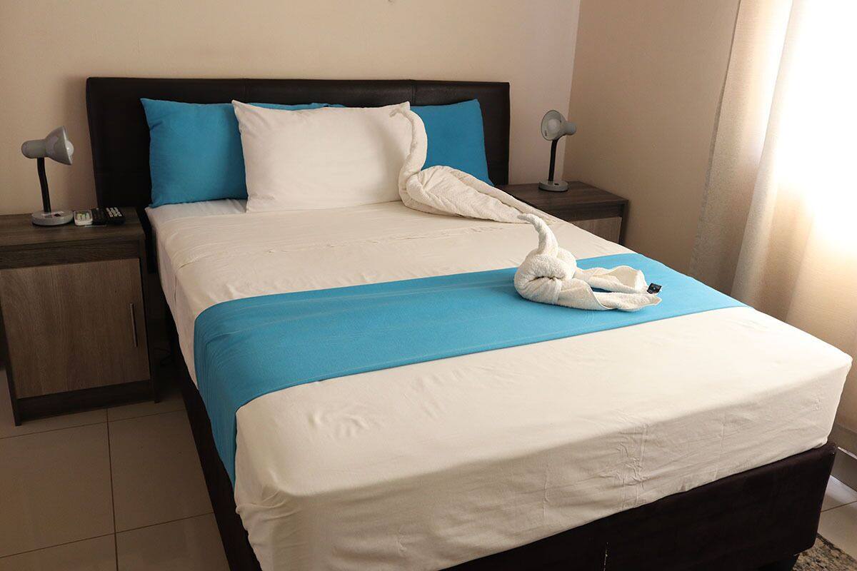 A neatly made bed with a blue bedspread and white pillows, a white towel shaped like a swan on the bed, and two bedside lamps on wooden nightstands.