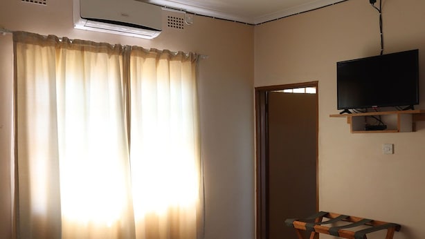 A hotel room with a bed, a TV mounted on the wall, and a window with curtains. - Amariah Guesthouse Newstance (Kasane)