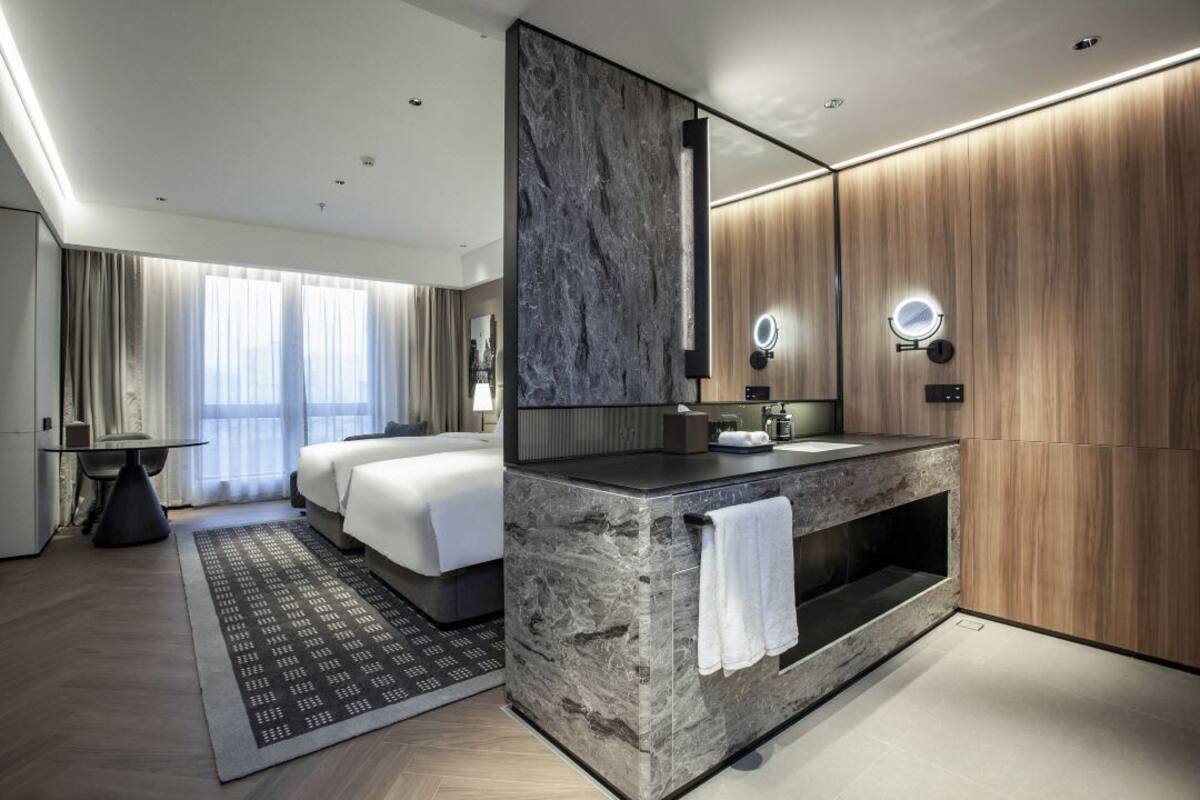 Quarto de hotel moderno com uma cama grande, uma toucador com espelho e um espaço de estar com mesa e cadeiras.