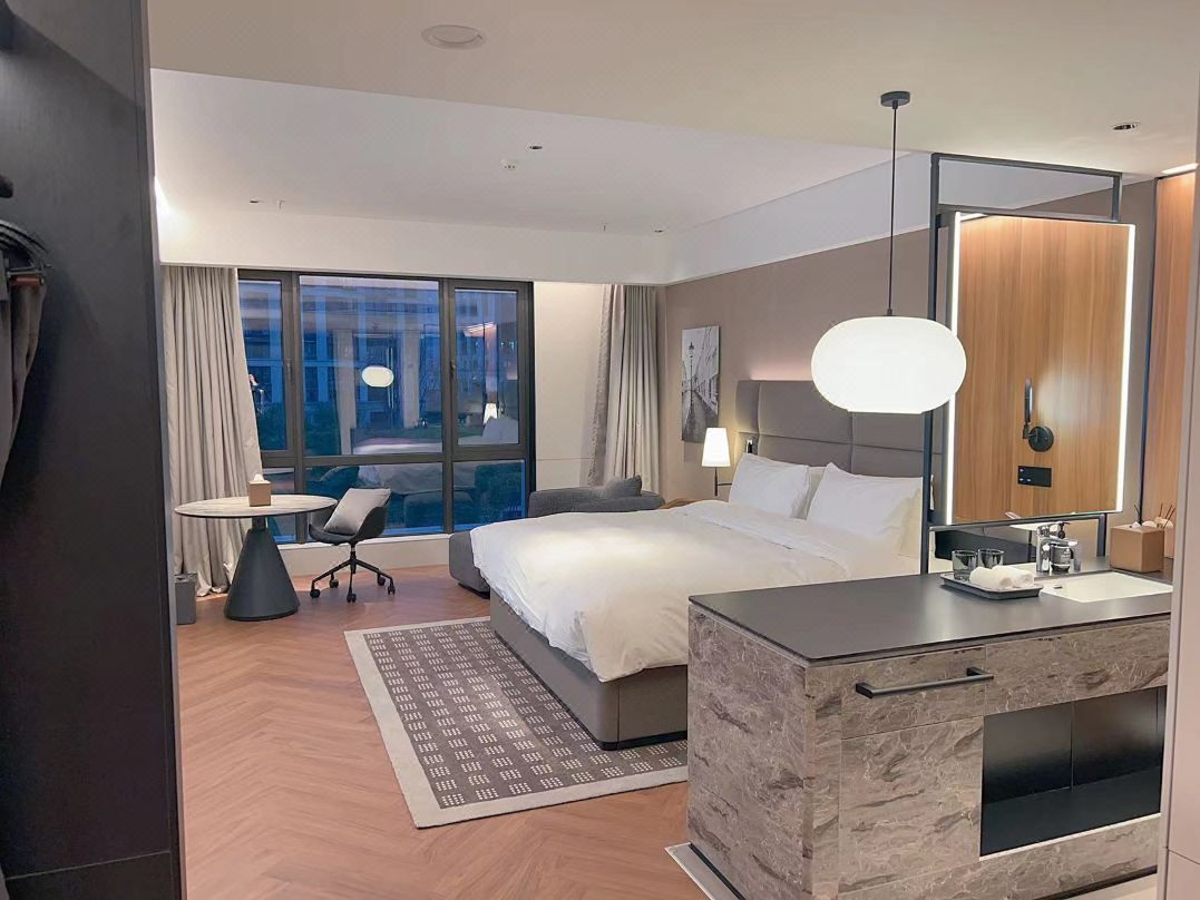 A modern hotel room with a large bed, a desk with a chair, a small table, a large mirror, and a view of the city.