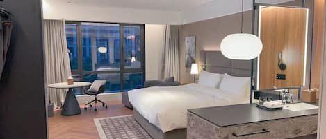 A modern hotel room with a large bed, a desk with a chair, a small table, a large mirror, and a view of the city.