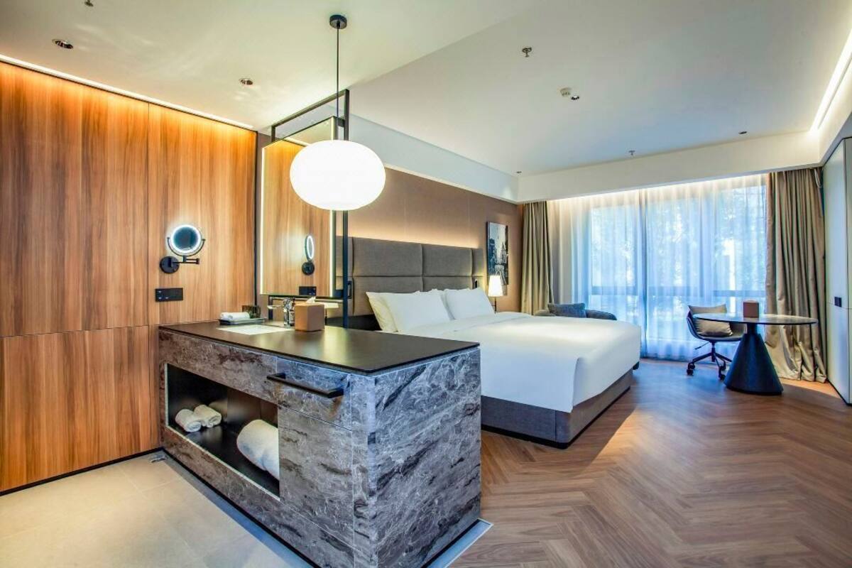 Quarto de hotel moderno com toucador de mármore, uma cama grande, uma mesa redonda e vista para a cidade pela janela.