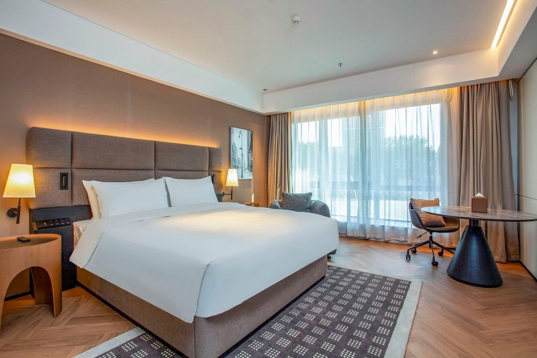 Habitación de hotel con una cama grande, mesitas de noche con lámparas, una pequeña mesa redonda, una silla y un amplio ventanal con cortinas.