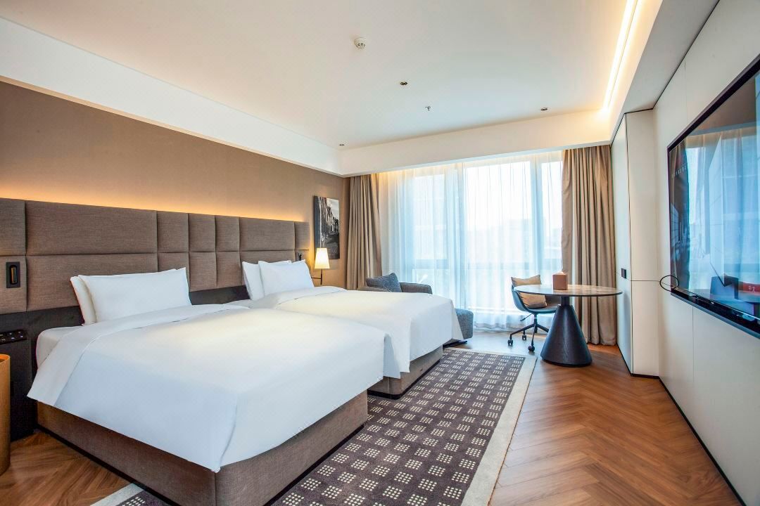 Habitación de hotel con dos camas, un escritorio, una silla y un ventanal con cortinas.