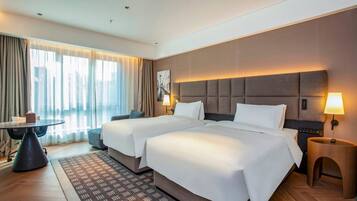 Habitación de hotel con dos camas, una mesita, una silla, una lámpara y vista a la ciudad.