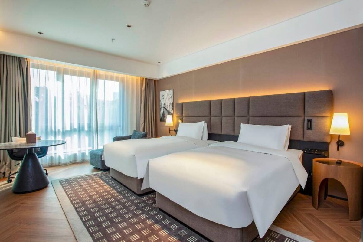 Quarto de hotel com duas camas, uma mesa pequena, uma cadeira, um abajur e vista para a cidade.