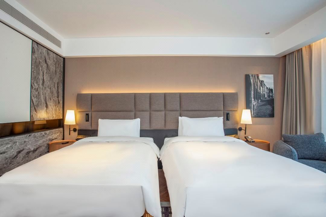 A hotel room with two beds, a headboard, bedside lamps, a sofa, and a painting on the wall.
