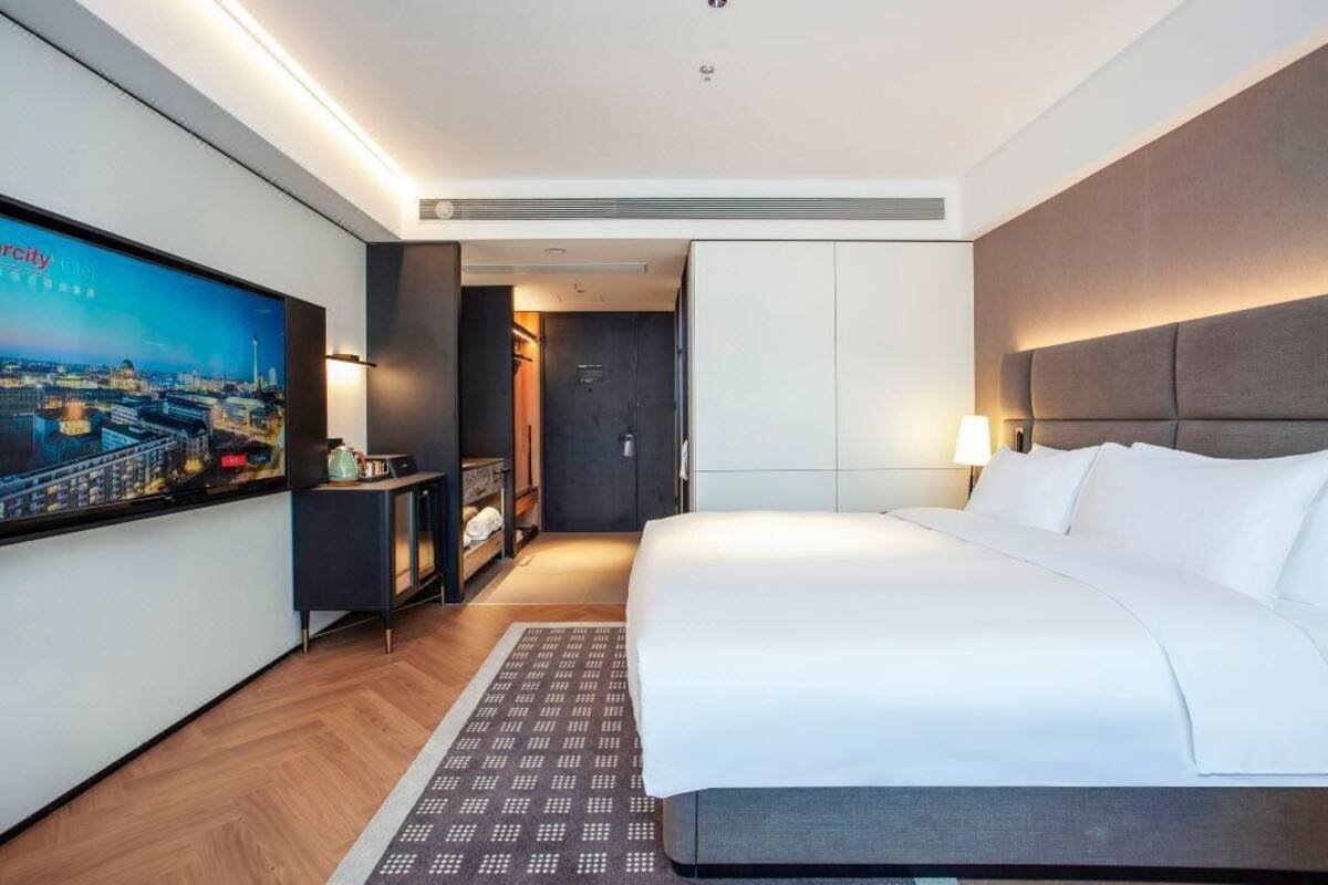 Quarto de hotel com uma cama grande, televisão na parede, guarda-roupa e uma mesa de trabalho com abajur.
