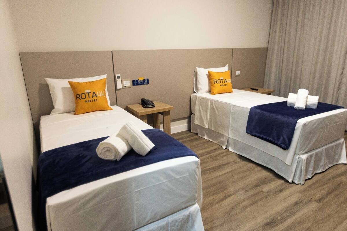 A hotel room with two beds, each with a white bedspread, a blue blanket, and a pillow with the hotel's logo. There is a small table with a phone and a sign indicating amenities.