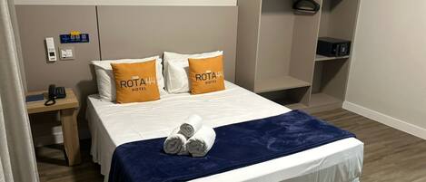 Una habitación de hotel con una cama, dos almohadas con el nombre del hotel, una mesita de noche con un teléfono y un armario.