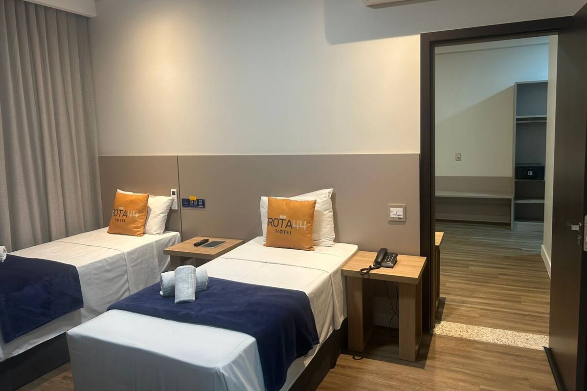 A hotel room with two beds, each with a pillow labeled "ROTA444 HOTEL". There is a bedside table with a phone and a remote control. The room has a wooden floor and a door leading to a hallway.