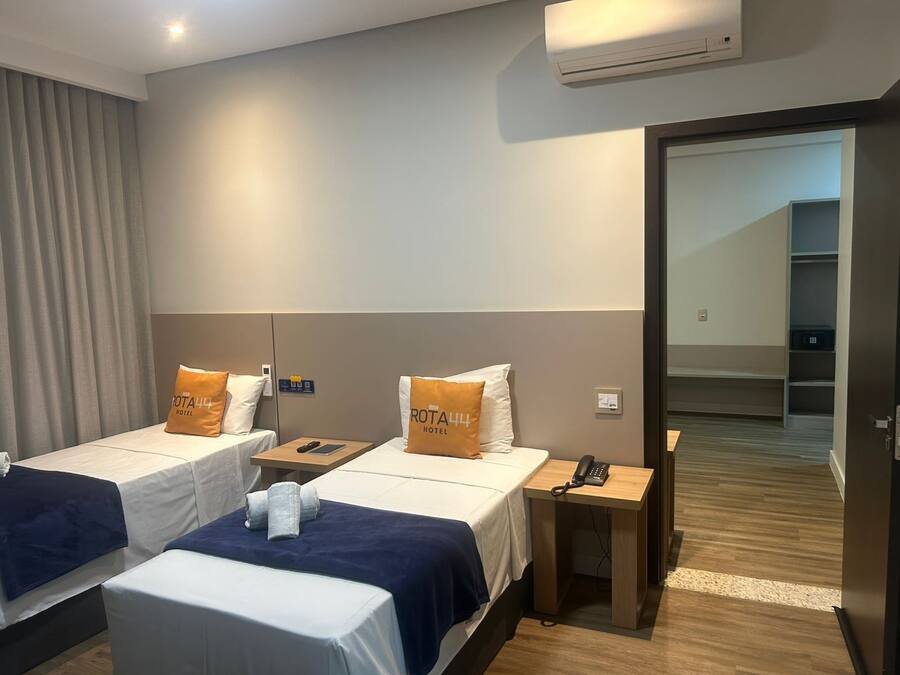 A hotel room with two beds, each with a pillow labeled "ROTA444 HOTEL". There is a bedside table with a phone and a remote control. The room has a wooden floor and a door leading to a hallway.