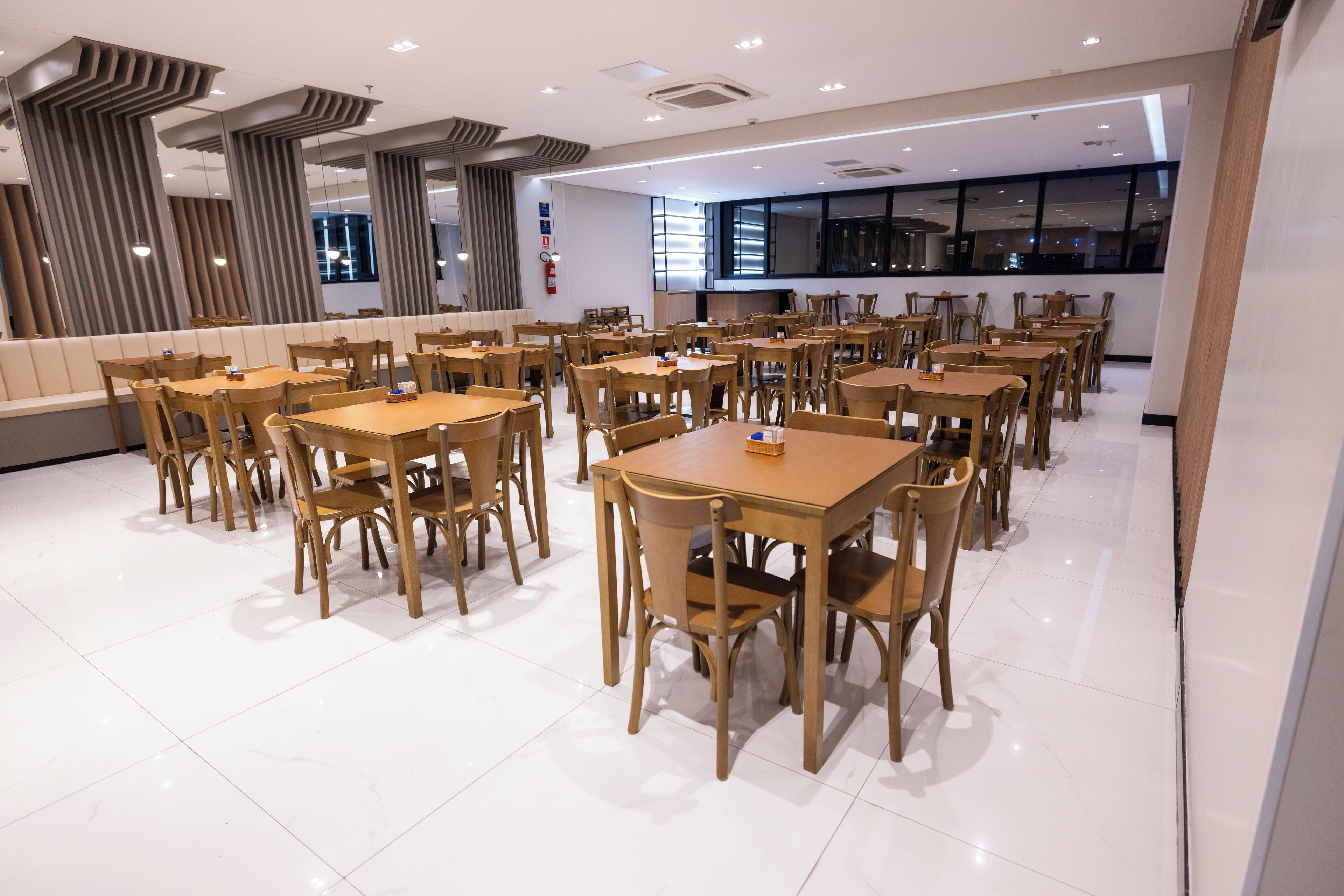 Un vaste espace dédié à la salle à manger, avec de nombreuses tables et chaises en bois, un design moderne au niveau du plafond et de grandes fenêtres qui laissent entrer la lumière naturelle.