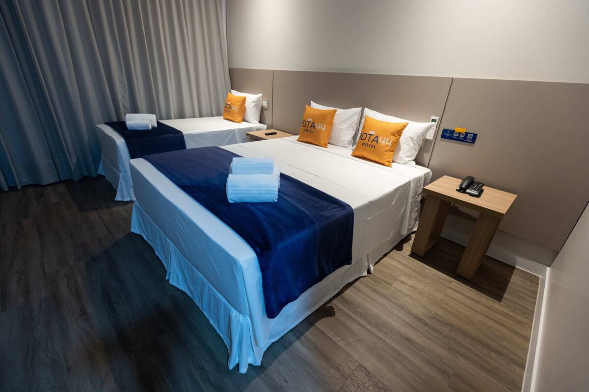 A hotel room with two beds, each with a blue and white bedspread, and orange pillows with the hotel name. There is a wooden nightstand with a phone and a sign indicating amenities.