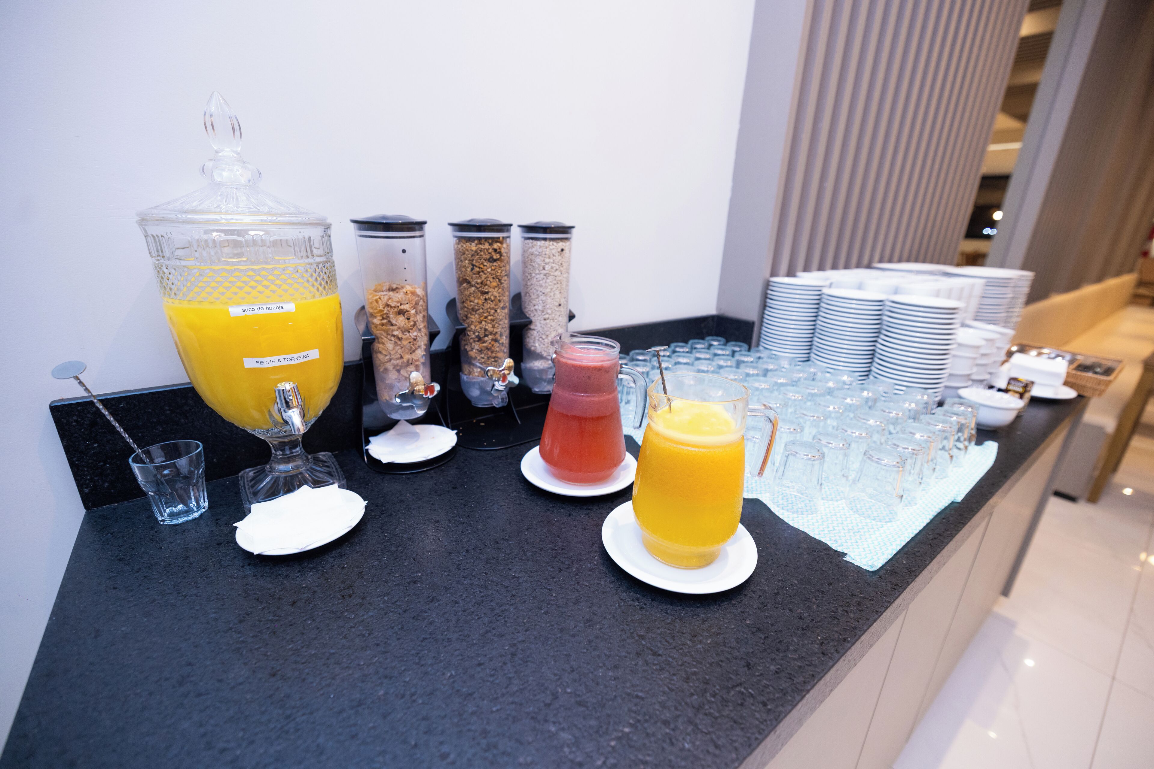A buffet setup with various food and drink options, including a large container of orange juice, cereal containers, and a glass of red smoothie.