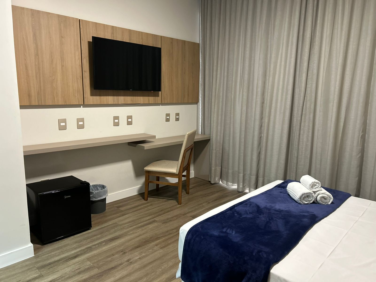 Une chambre d’hôtel avec un lit, un bureau, une chaise, une télévision et une petite valise noire.