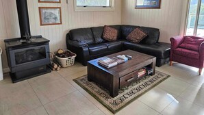 A living room with a black leather sofa, a wooden coffee table, a black wood-burning stove, and a large window with a view of trees.