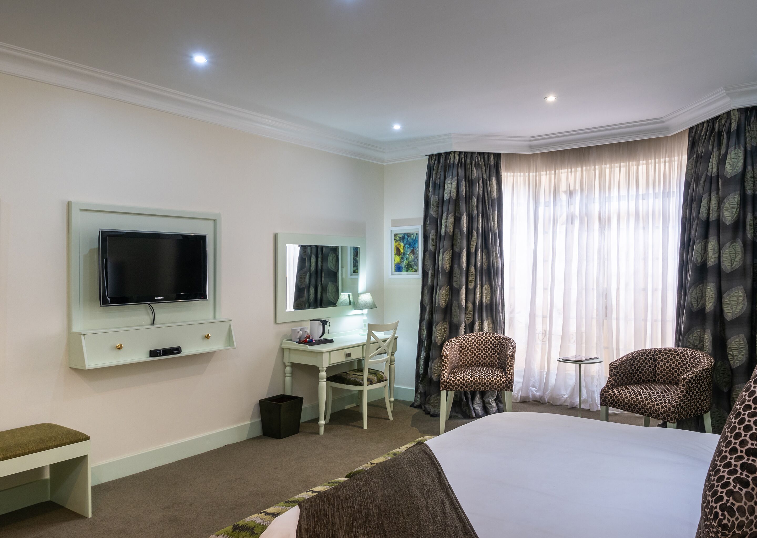A hotel room with a bed, a desk with a chair, a television, a mirror, and a window with curtains.