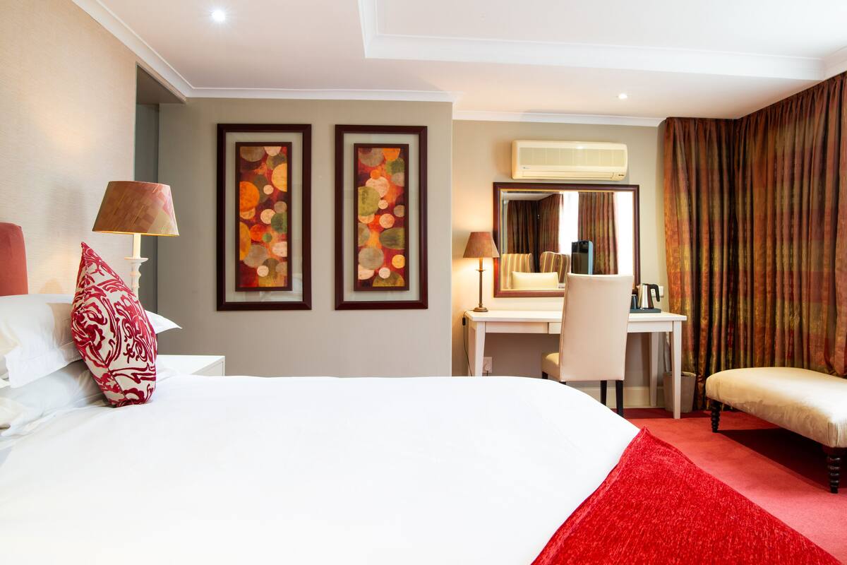 A hotel room with a large bed, a desk, a chair, and a red carpet. There are two framed artworks on the wall and an air conditioning unit above the desk.