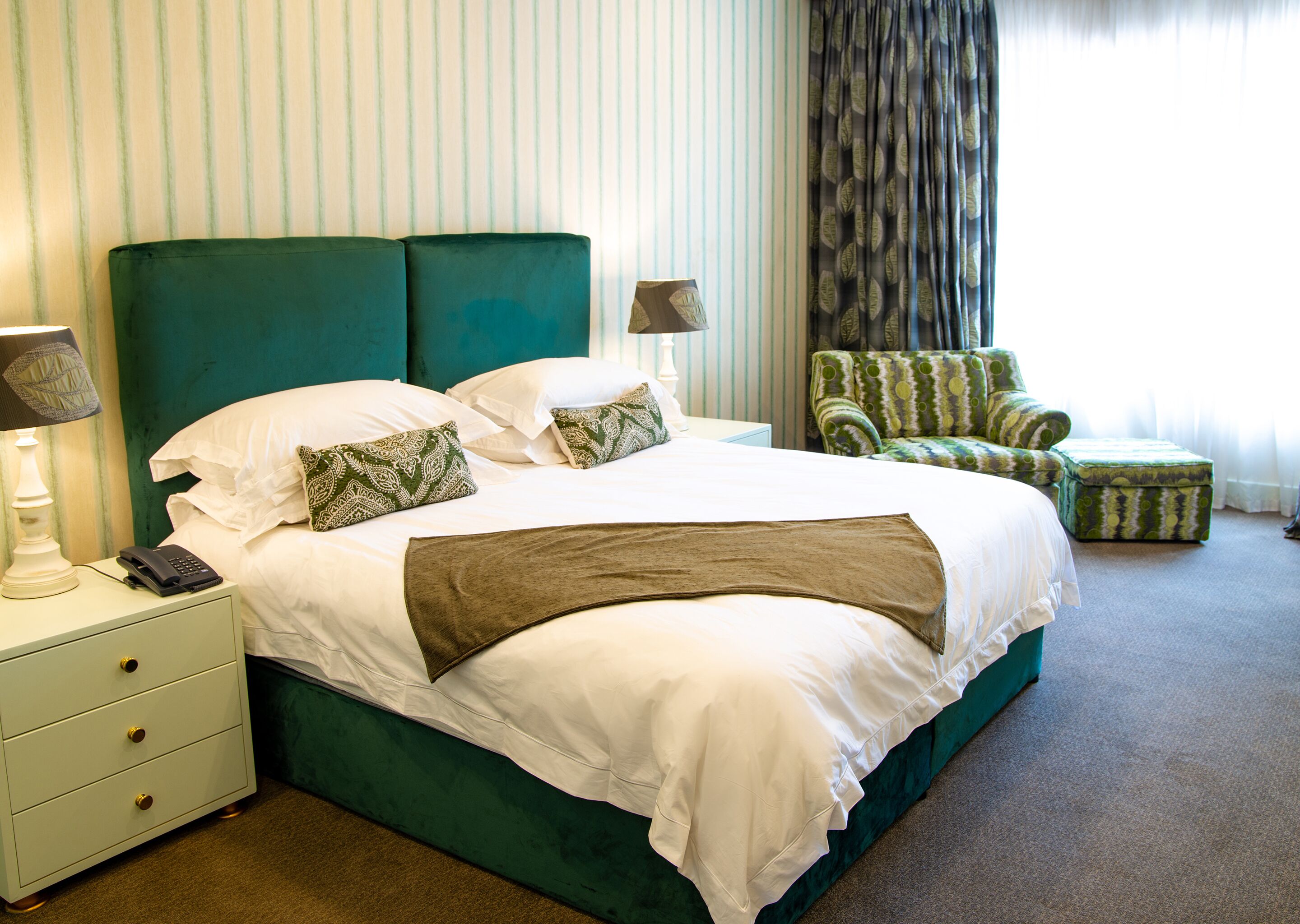 A hotel room with a large bed, a nightstand with a lamp, a chair, and a small ottoman.